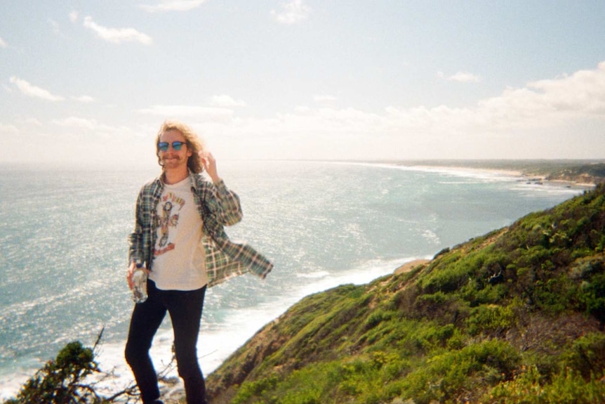 A young man with long curly hair and sunglasses stands on the edge of a cliff