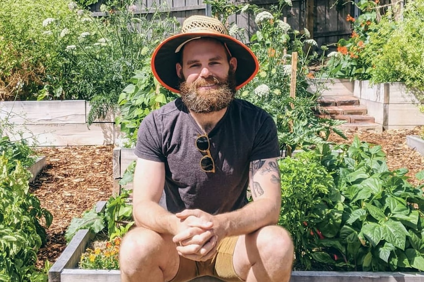A man sits on the edge of a raised garden bed in his Hobart garden. He grows food for himself and to share.