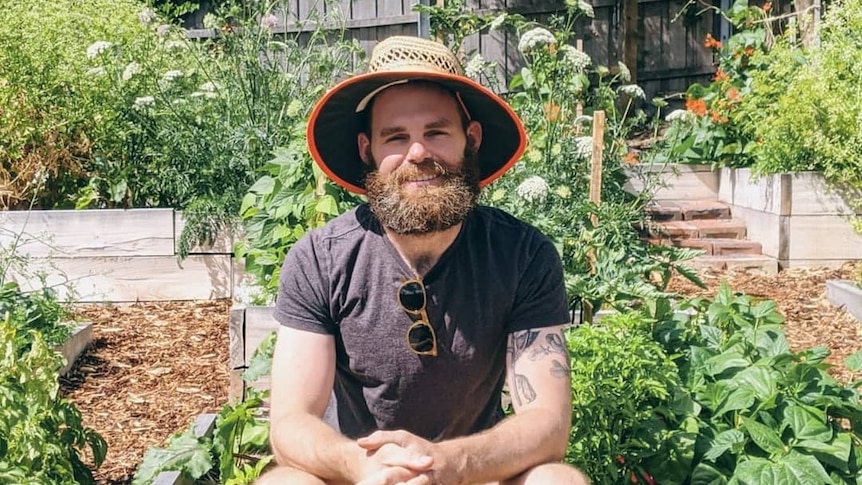 A man sits on the edge of a raised garden bed in his Hobart garden. He grows food for himself and to share.