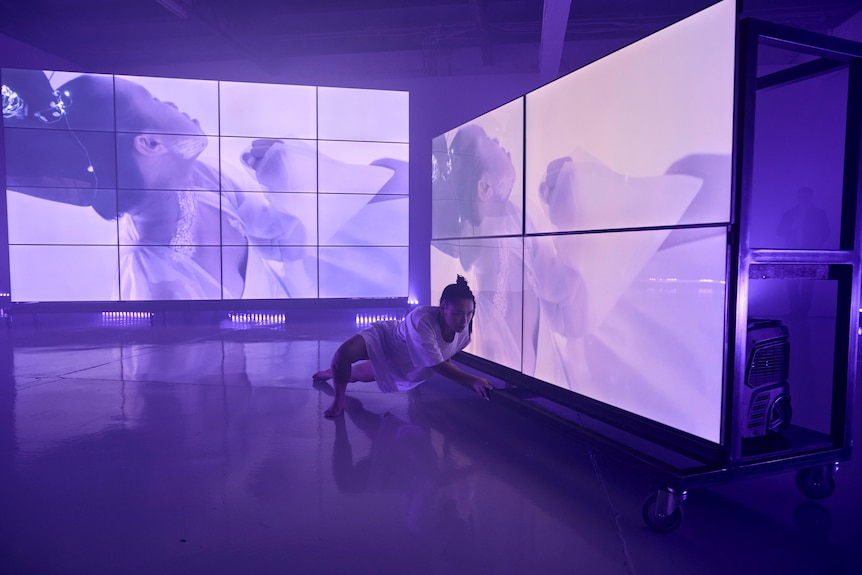 A dancer in all white is crouched on the floor in front of two large projection screens and purple lights.