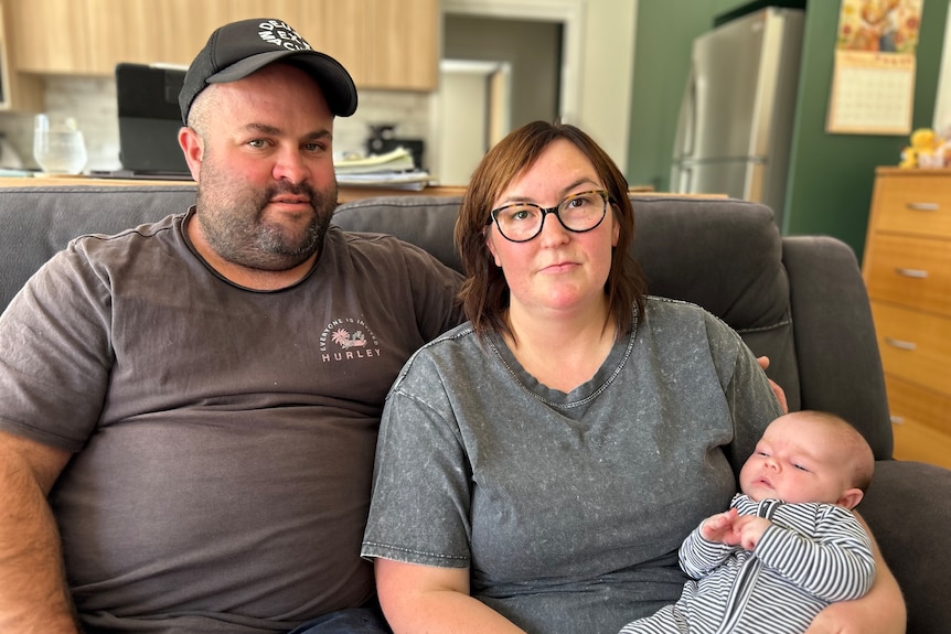 A man and a woman sit on a couch, looking seriously at the camera. The woman is cradling a baby.