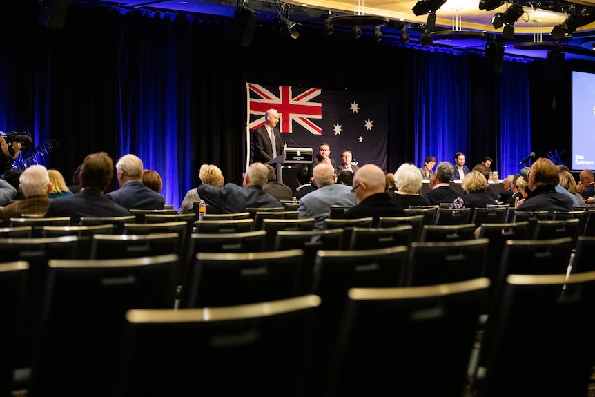 A man stands at a lecture with a crowd in front and empty chairs in the foreground.