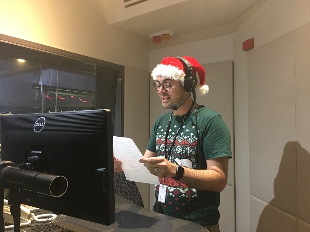 A man stands in a radio studio holding a piece of paper dressed in Christmas attire