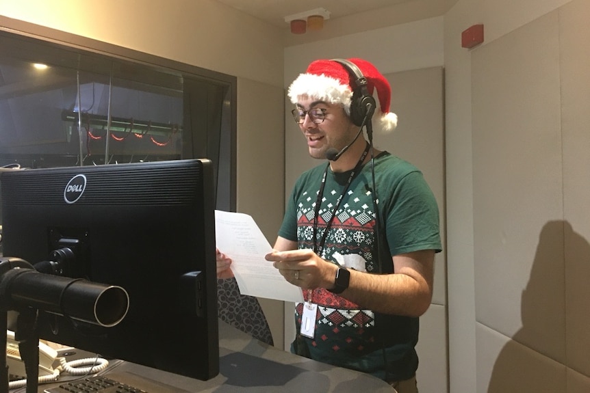 A man stands in a radio studio holding a piece of paper dressed in Christmas attire