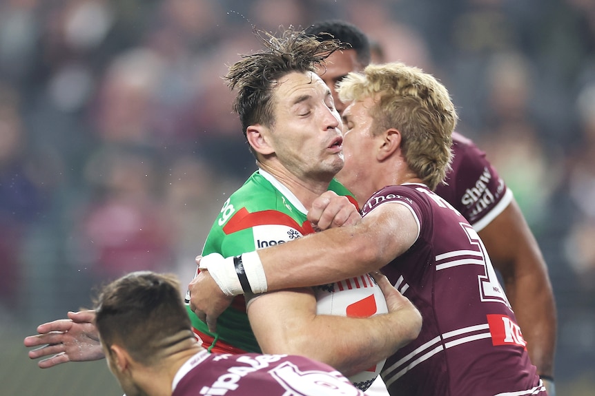 A South Sydney NRL player grimaces as he is holds on to the ball while being tackled.