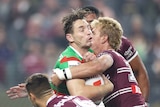 A South Sydney NRL player grimaces as he is holds on to the ball while being tackled.