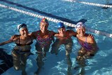Four smiling women in swimming caps and goggles with their arms around each other in a pool.