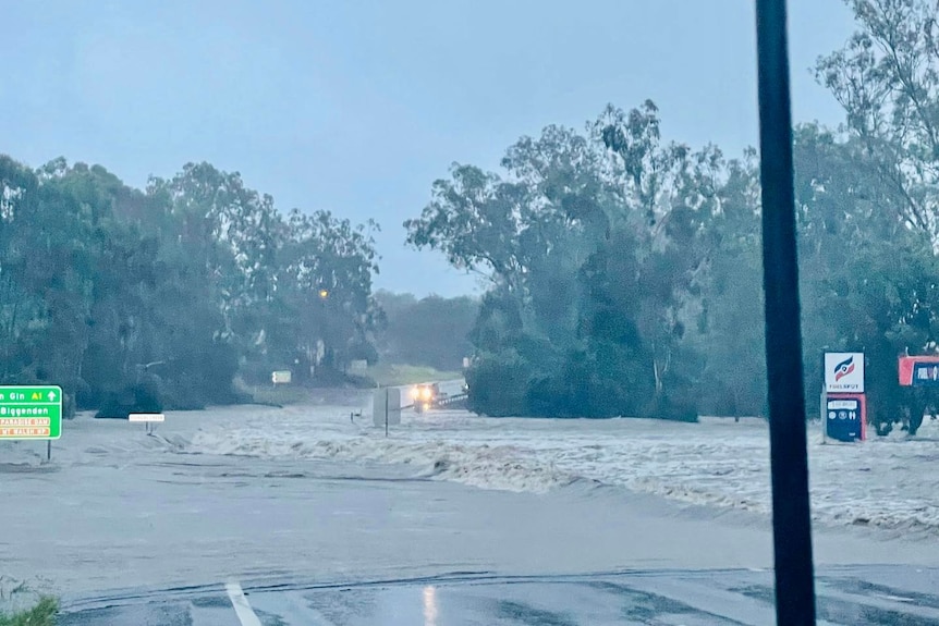 A torrent of brown floodwater sweeps across a regional queensland road beside a fuel station 