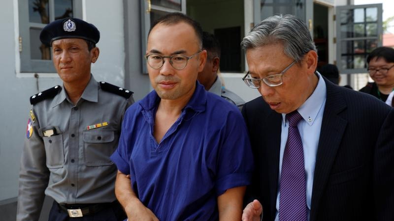 A man in prison uniform is flanked by a guard and another man.