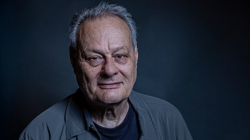 Artist Mike Parr wears a grey jacket. He poses with a kind expression in front of dark grey background.