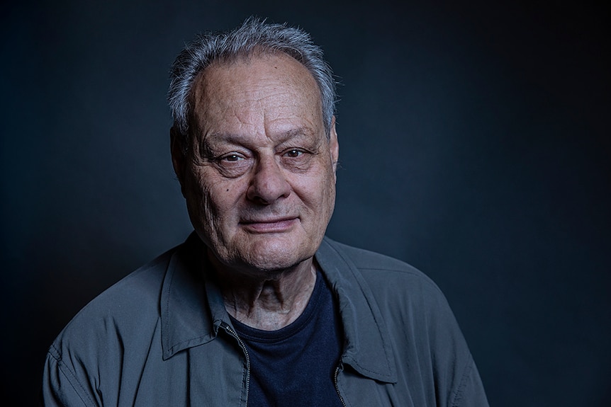 Artist Mike Parr wears a grey jacket. He poses with a kind expression in front of dark grey background.