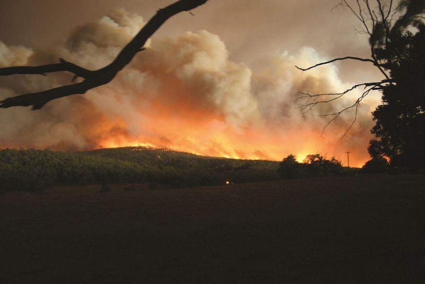 A large plume of smoke hovers over the mountains as a bushfire rages, sending out orange flames