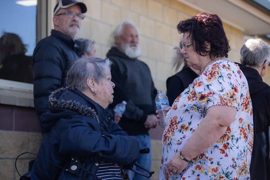 Une dame âgée assise parle à une femme plus jeune parmi une foule de personnes devant un centre médical.