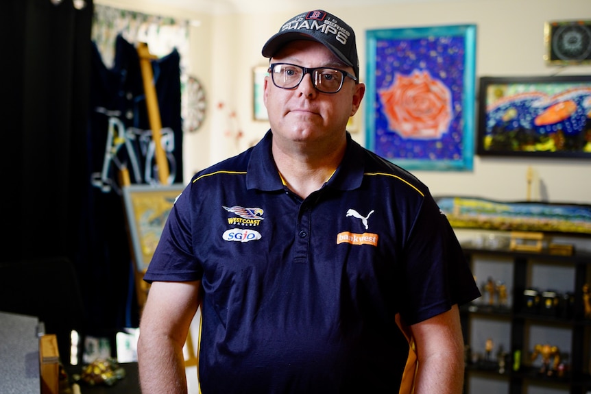 Ray Smith stands in a room with artwork behind him. He looks seriously at the camera.