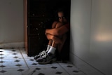 A child sits on the floor hunched in a corner next to a cupboard, peering over their knees.