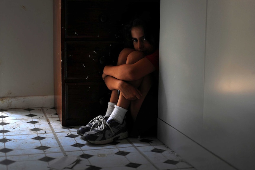 A child sits on the floor hunched in a corner next to a cupboard, peering over their knees.