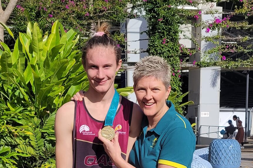 Young boy with hair tied in pink band with Natalie Cook. Green shurbs, flowers, white building behind. Cook holds medal.