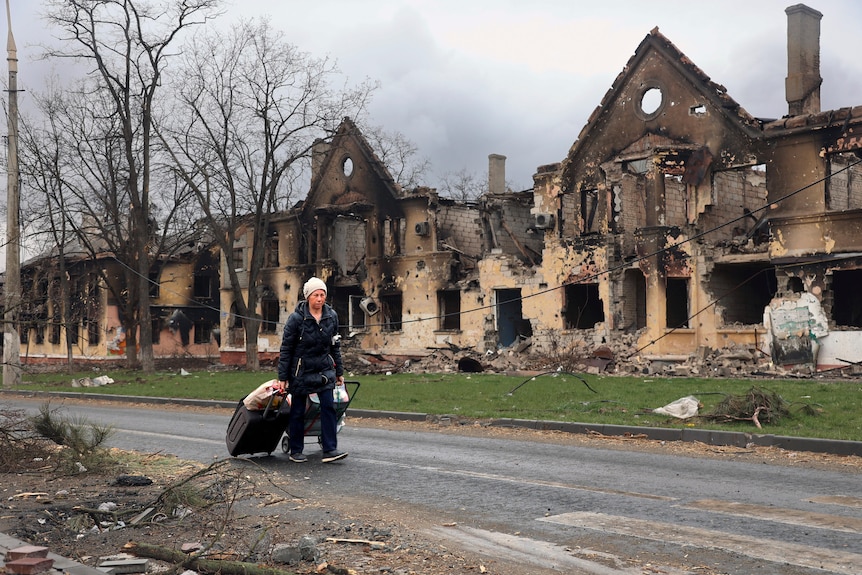 A woman walks down a road pulling bags behind her in front of houses reduced to rubble by bombings