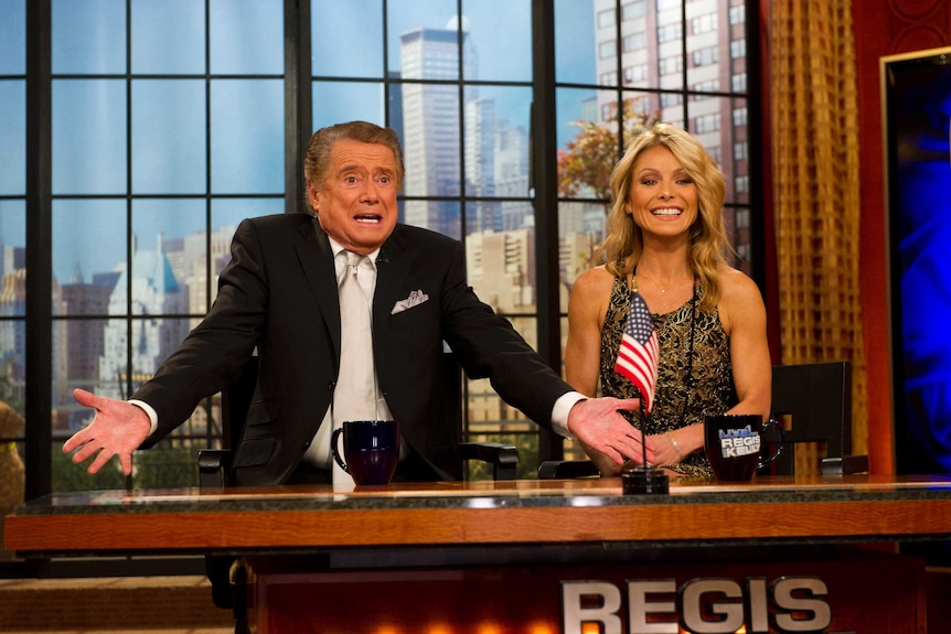 An elderly man with an animated expression and arms in front of him sits next to a laughing woman.