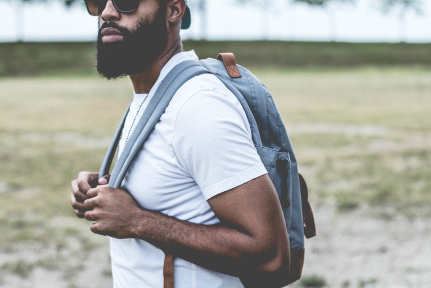 A man wearing a white t-shirt holds on the straps of a backpack.