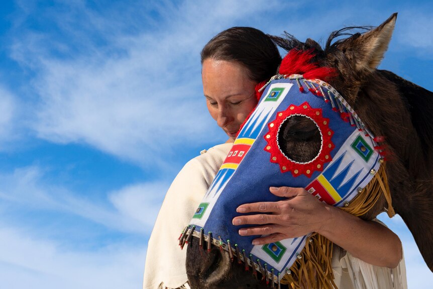 A woman embraces a horse which is wearing traditional Native American head regalia  