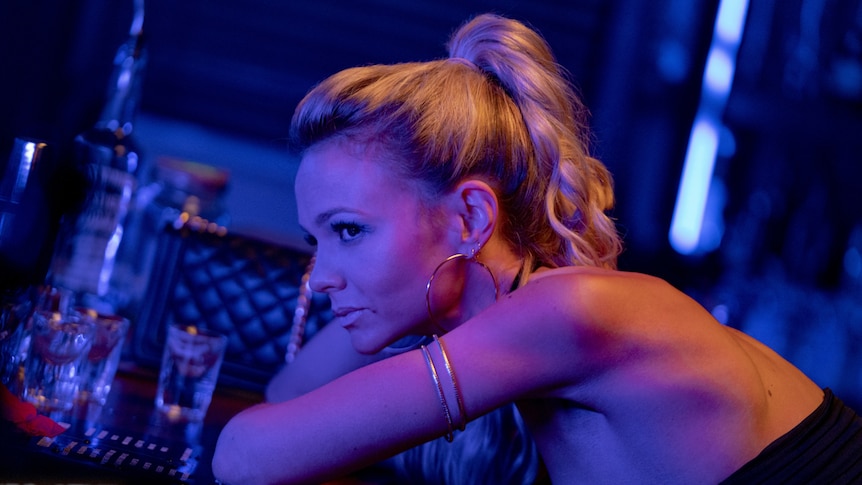 Blue-lit darkened interior shot in bar with young woman with long light blonde hair in ponytail leaning on bar.
