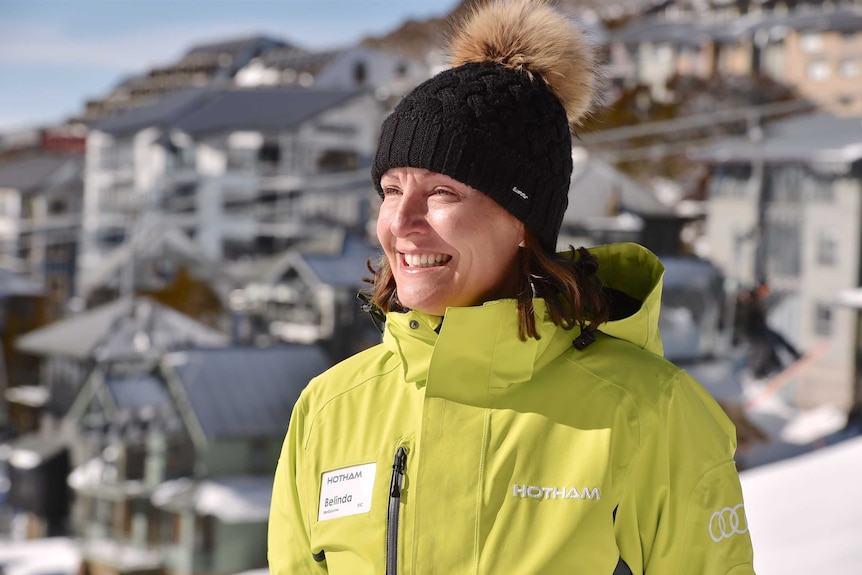 A woman in a yellow snow jacket, wearing a black beanie, standing in front of an alpine resort.