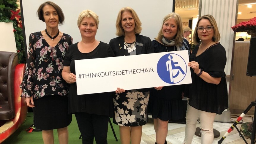 Five women stand holding a sign saying that says 'think outside the chair' with a hashtag symbol.