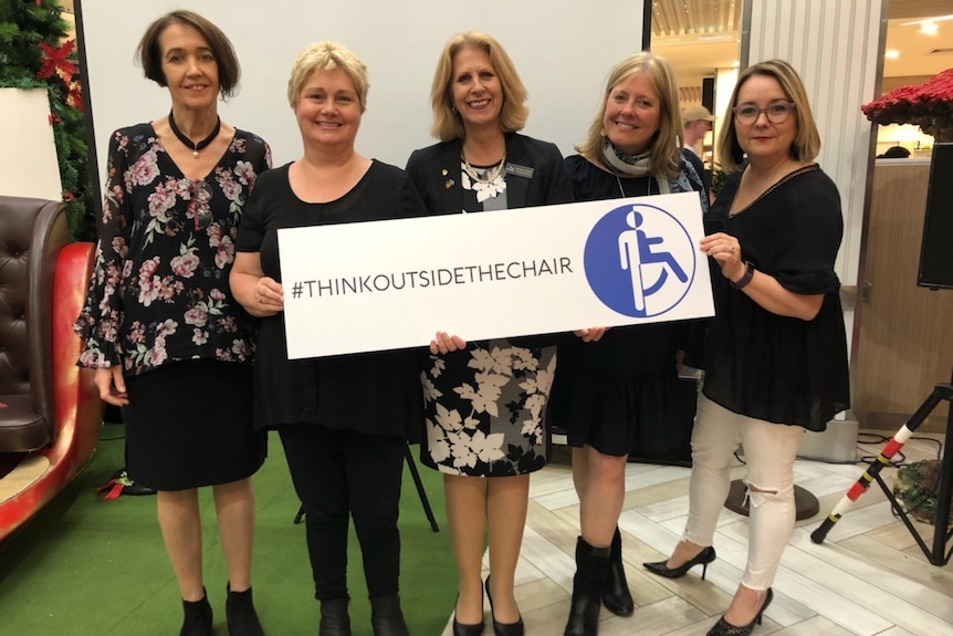 Five women stand holding a sign saying that says 'think outside the chair' with a hashtag symbol.