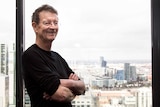 A man stands with his arms crossed, smiling, next to a window overlooking a city.