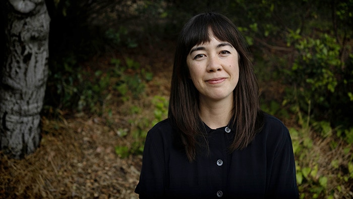 Jenny Odell, with shoulder-length black hair and black shirt, sits before blurred greenery behind her, smiling widely.