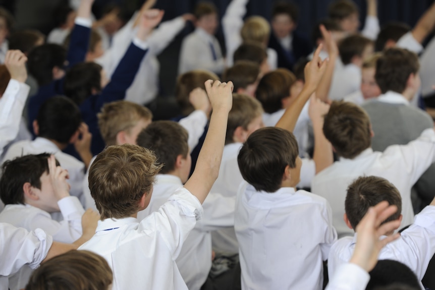 The back of teen boys' heads at a school assembly with a few with their hands up.