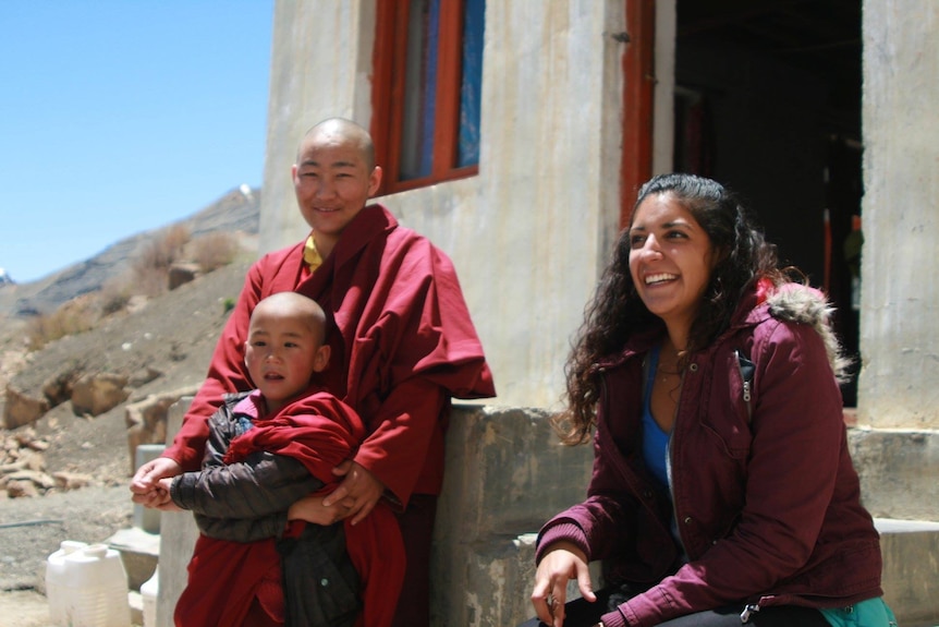 Une femme est assise à l'extérieur d'un bâtiment en béton avec un moine tibétain et un enfant.