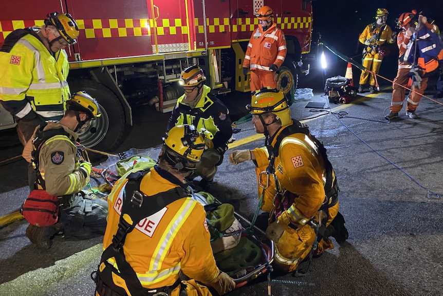 Men in CFA uniforms with a stretcher, kneel on road