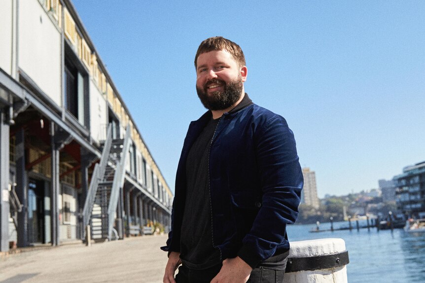 Kip Williams, a man in his early 40s, smiles, leaning against a structure on an outdoor wharf.