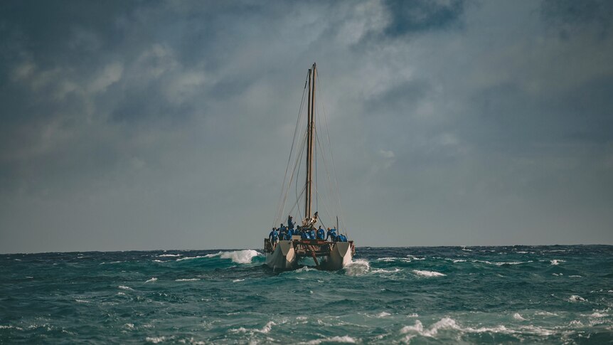 The vaka sails off into the distance with small waves pushing up against it. 