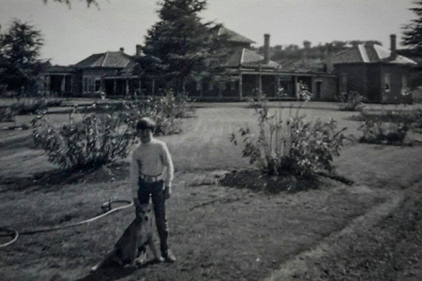 Black and white image of a young boy holding a dog by the collar poses in front of a country homestead