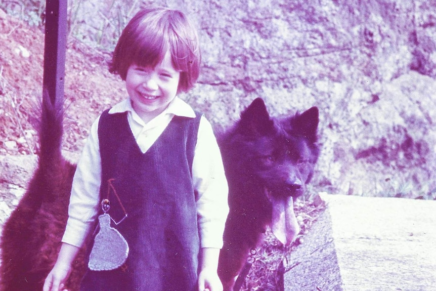 A female child smiles at the camera with a big black dog behind her.