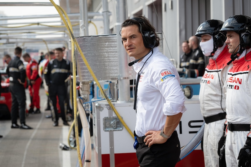 Orlando Bloom standing in the pits at a motor race with a concerned expression.