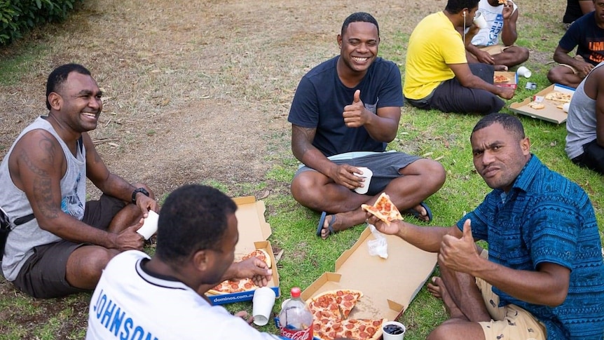 A group of Fjijian men sit on the grass eating pizza. 