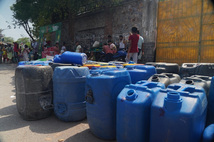 A dozen emptry water barrels and containers lined up with people standing in the shade.