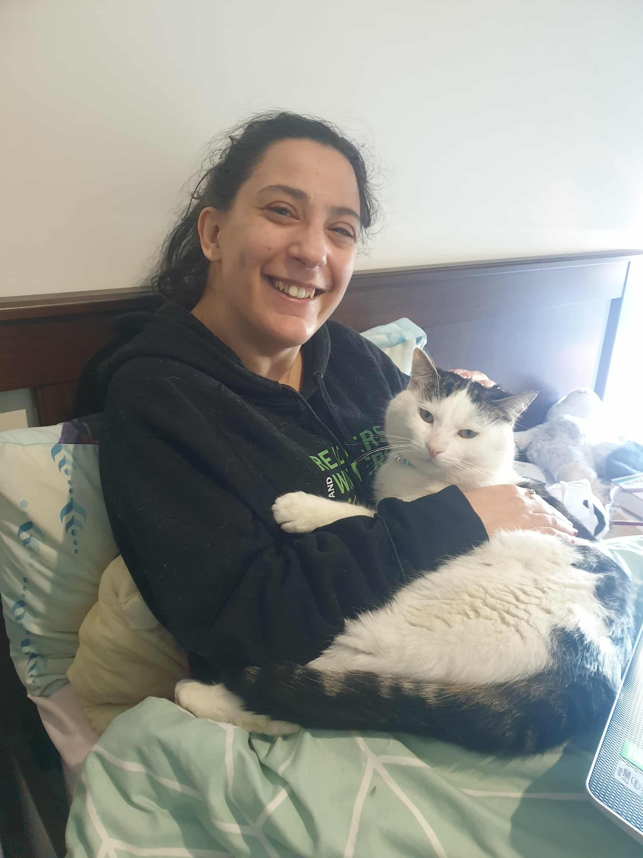 Laura, que tem longos cabelos escuros, está sentada na cama embalando seu gato Giles, um grande gato malhado preto e branco.