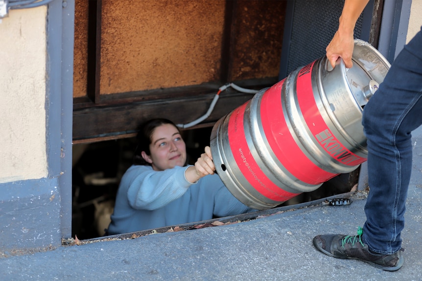 A woman in grey jumper is handed a red and silver keg into a cellar