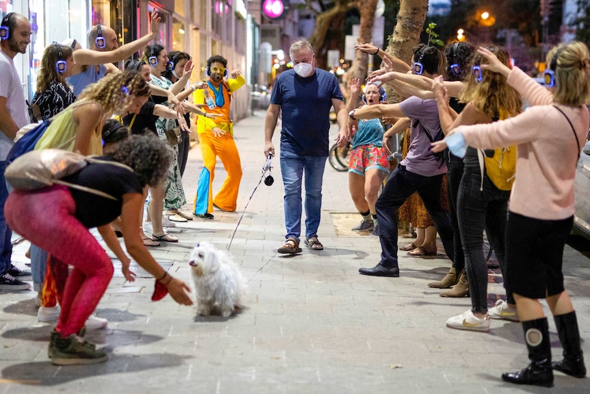 Un grup de oameni care poartă căști făcând o cale pentru un bărbat purtând o mască de față plimbându-se cu un câine