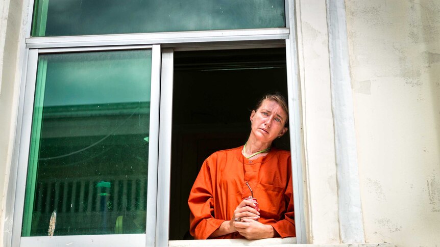 A woman in prison oranges stands at an open window with a distraught look on her face