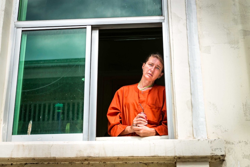 A woman in prison oranges stands at an open window with a distraught look on her face
