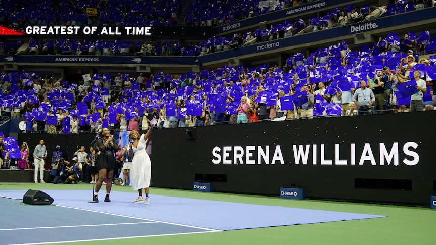 Serena Williams on court at the US Open waving at the crowd, with signs saying 'greatest of all time' and 'Serena Williams'.