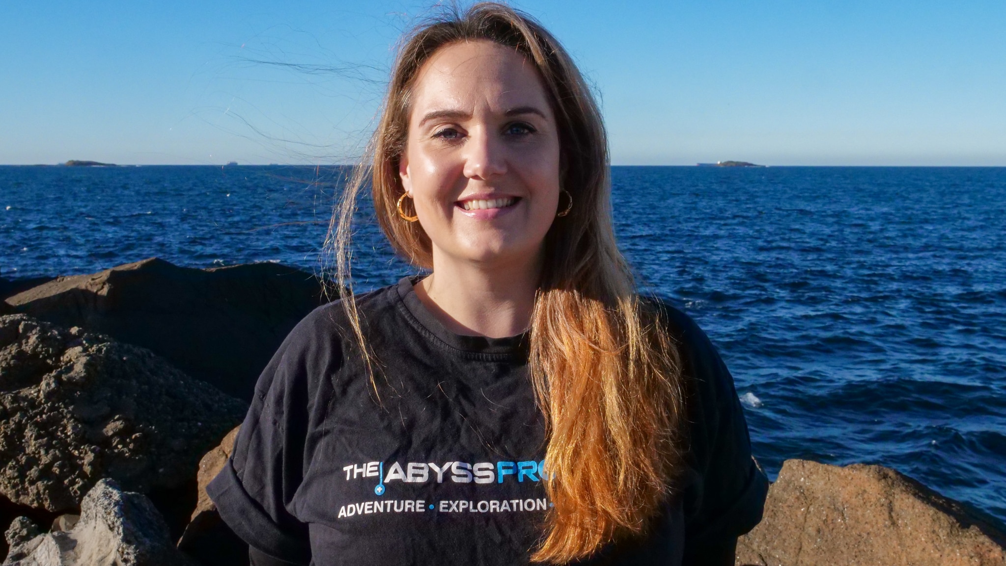 Uma mulher de cabelos castanhos está em um píer com ilhas ao fundo. 