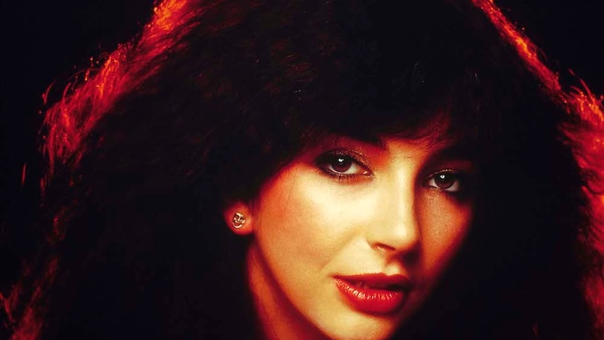 Close up photograph of Kate Bush, her brown hair is styled big around her face, the back ground is black with a red light behind