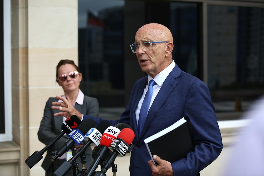 A man in a navy blue suit speaks at a press conference as a woman watches in the background.